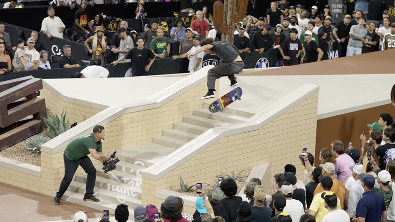Camera operator capturing live skate competition action, photographer visible at the base of the ramp